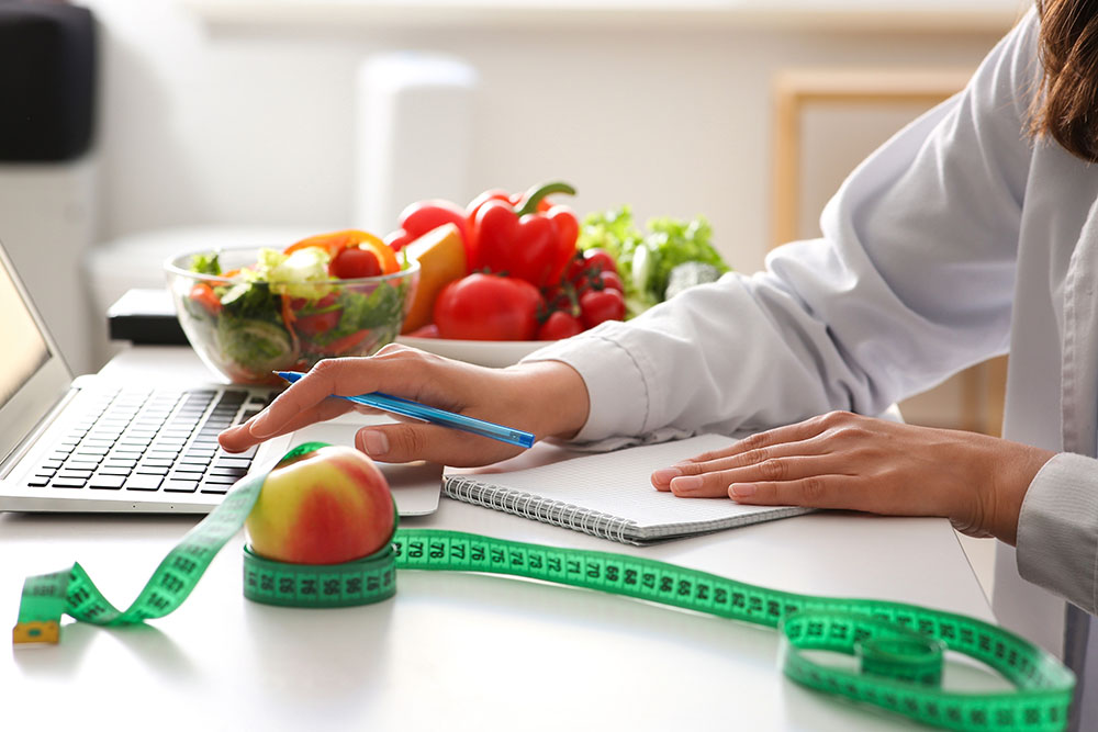 Young female nutritionist taking notes and using a laptop at her office desk.